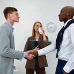 Professional meeting with two men shaking hands and a woman with a coffee cup in an office.