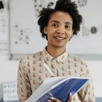 Confident female teacher smiling while holding a notebook in a classroom setting.