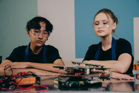 Two young engineers wearing safety goggles focus on an electronic project indoors.